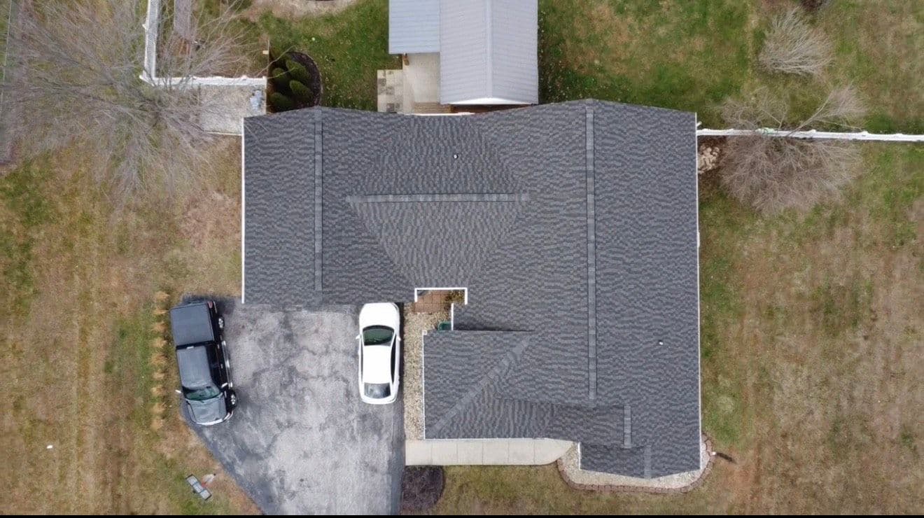 Aerial view of a house with a gray roof and driveway, surrounded by grass and trees.